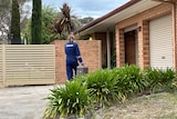 A woman in blue suit with "forensic" written on the back of it walks outside a suburban brick home
