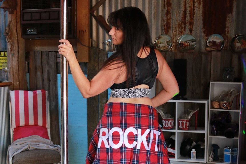 A woman standing next to a pole in a shed where's a flannelette jacket around her waist emblazoned with the word 'rockit'.