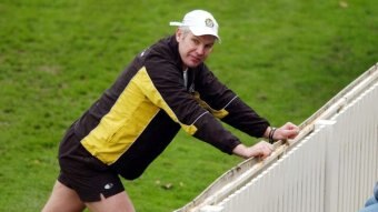 Man wearing wearing shorts, long sleeve top and a white cap leans into a fence around a field with his arms outstretched.