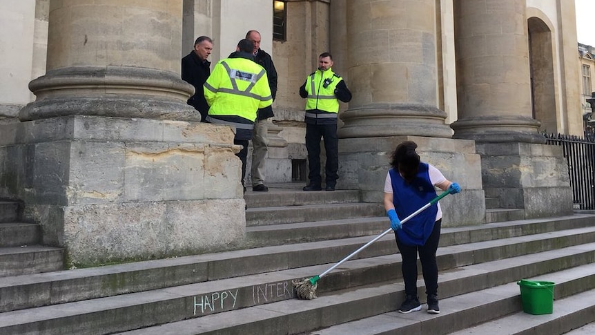 Female cleaner mops away message written on steps as a group of men converse at the top.