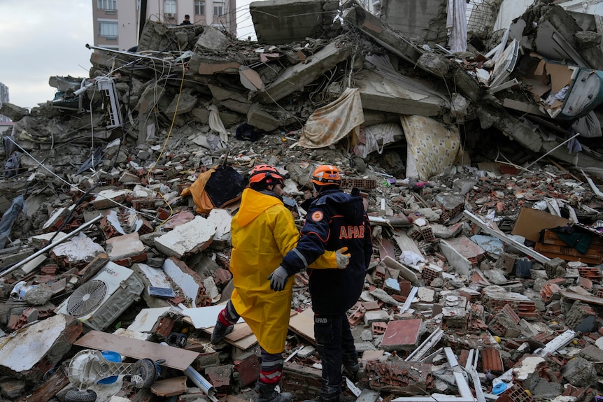 Two workers in uniform stand with their arms around one another looking at the rubble from that surrounds them. 