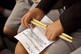A boy holds a sheet of music and a pair of drumsticks on his lap.