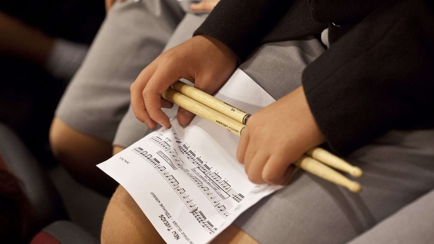 A boy holds a sheet of music and a pair of drumsticks on his lap.