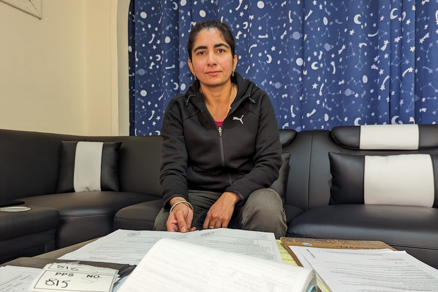 A Punjabi-Australian woman, Raj Ghuman, sits on a leather couch handling paperwork in front of blue starry curtains.