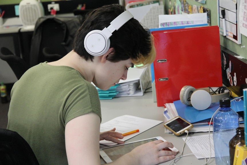 Sage Nikolovski completes work on an ipad in a classroom while wearing white headphones.