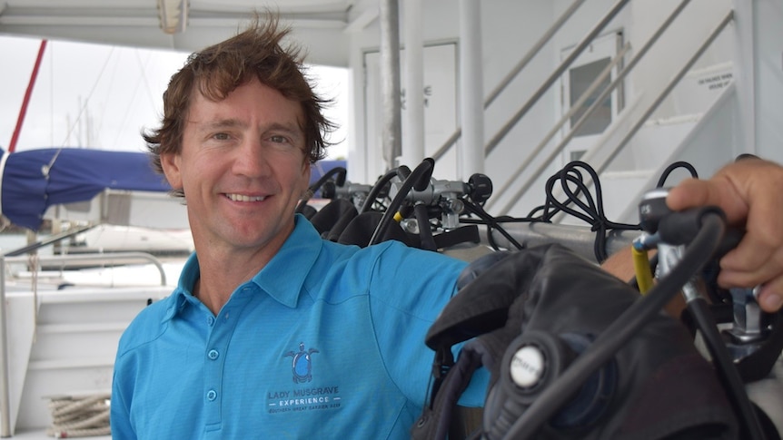 A man in a blue polo shirt smiles at the camera, and sits next to a row of diving gear and equipment on a boat.