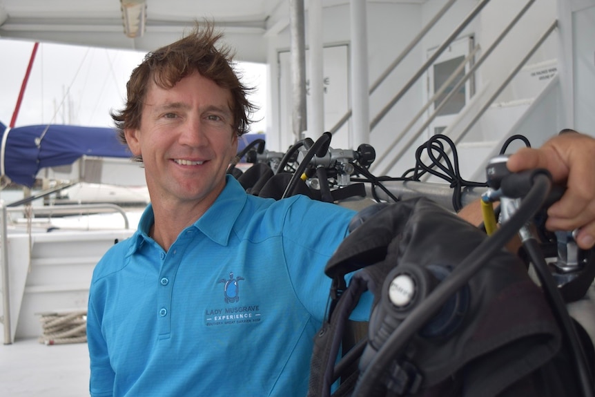 A man in a blue polo shirt smiles at the camera, and sits next to a row of diving gear and equipment on a boat.