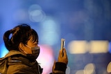A woman wears a mask and holds a smartphone up.