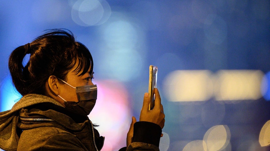 A woman wears a mask and holds a smartphone up.