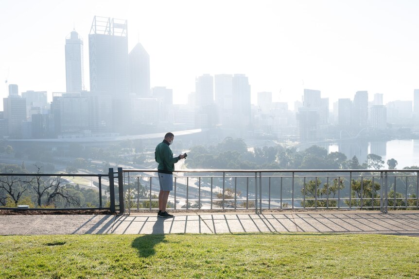 Man wearing mask and active wear checks his phone with city skyline in background. 