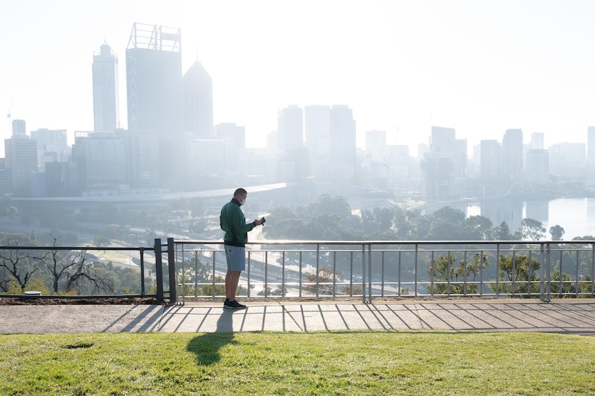 Man wearing mask and active wear checks his phone with city skyline in background. 