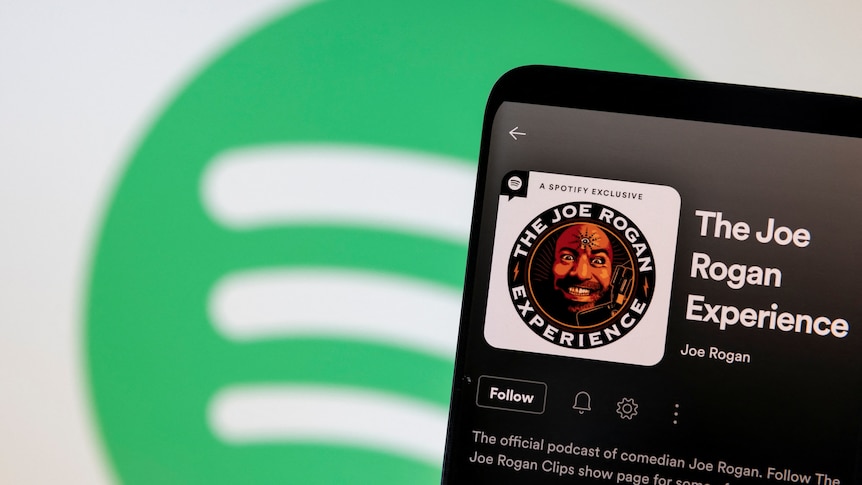 A phone showing a logo for The Joe Rogan Experience podcast on the Spotify app is held in front of a Spotify logo