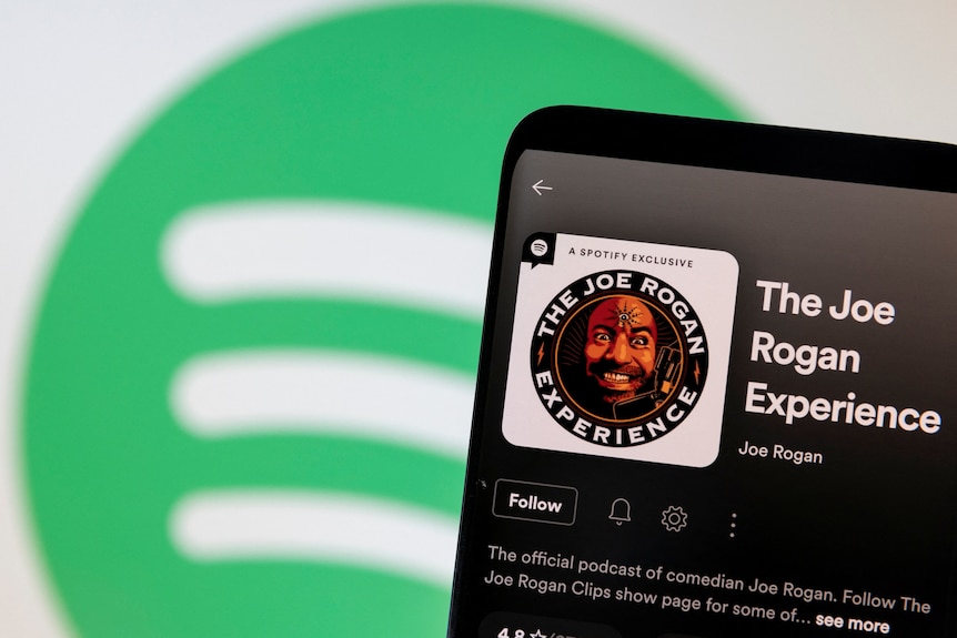 A phone showing a logo for The Joe Rogan Experience podcast on the Spotify app is held in front of a Spotify logo