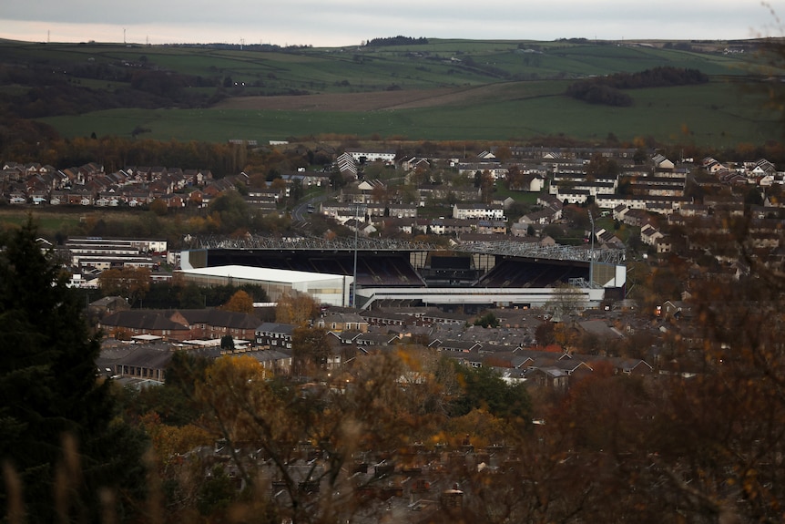 A wide-shot of a town dotted with houses and a big football stadium overlooking green hills.