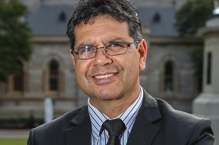 A man wearing a suit and glasses smiles at the camera standing outside a university