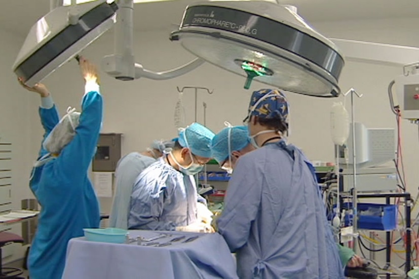 Four men and woman in surgical scrubs, blue head caps, white masks, goggles, perform on a patient, one adjusts a round light.