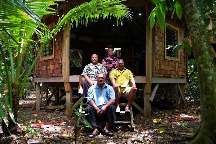 Three men and a woman sit on the steps outside a traditional Tongan building.