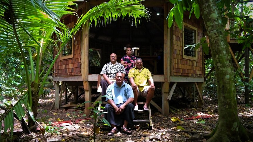 Three men and a woman sit on the steps outside a traditional Tongan building.