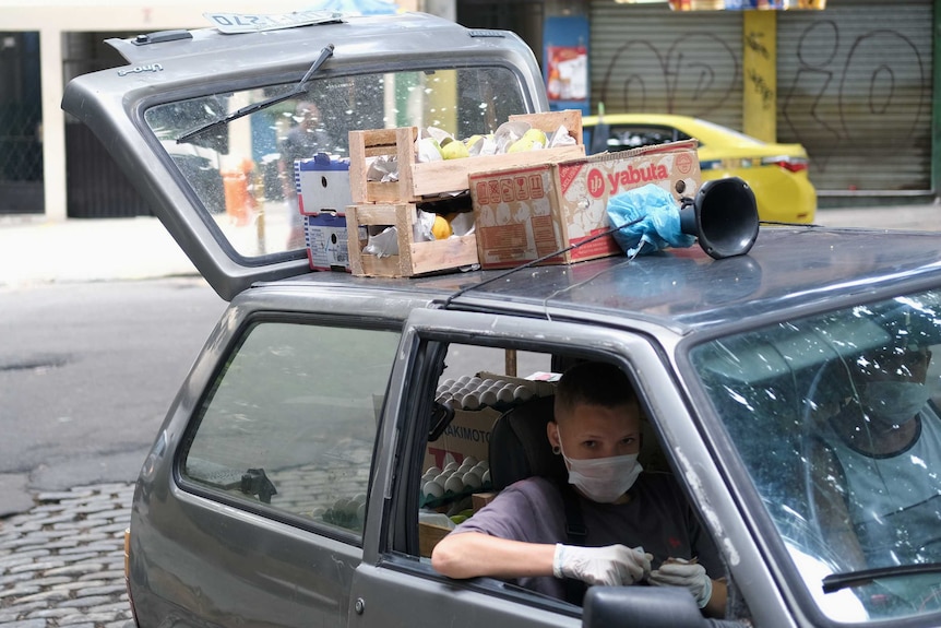 A person in a car wearing a face mask looks out the open window. On top of the care are crates of produce