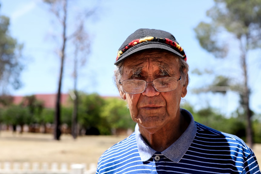 Man wearing black hat with glasses and blue shirt with trees and dry grass in background