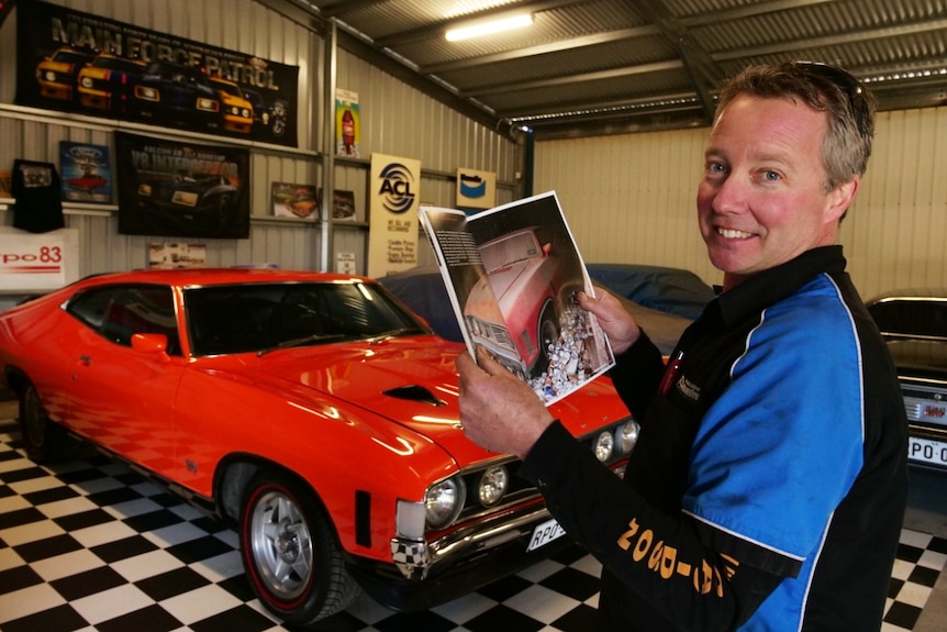 A smiling man looks at the a magazine displaying an old car, with a renovated similar car in the background.
