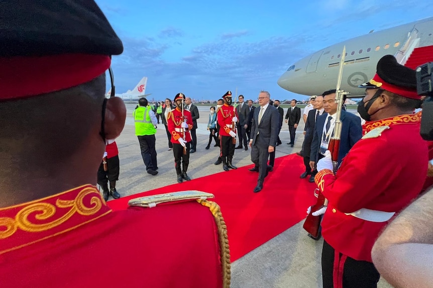 A man in a suit walks down a red carpet in front of a large grey plane, flanked by military personnel in red dress uniform.