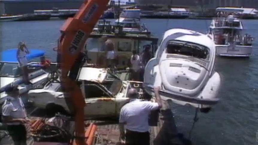A white VW is pulled out of a waterway by a red crane on a boat holding other cars