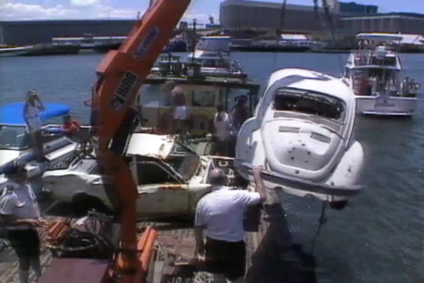 A white VW is pulled out of a waterway by a red crane on a boat holding other cars