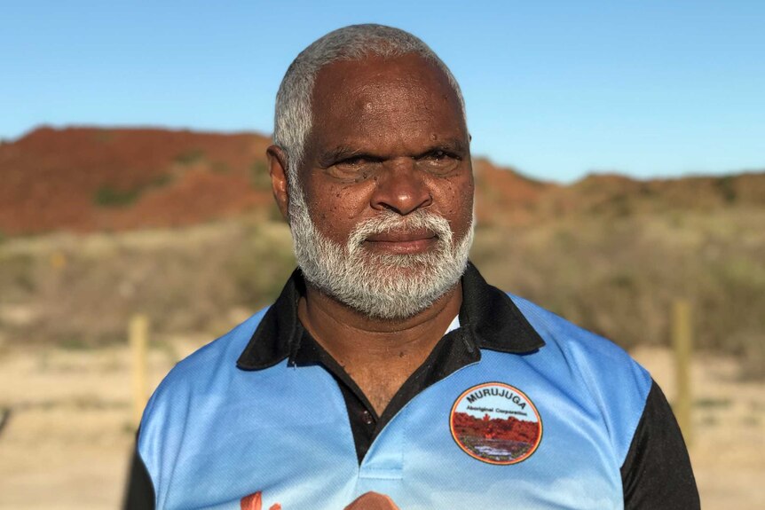 Head shot of an older Aboriginal man wearing a light blue shirt with red, rocky hills in the distance