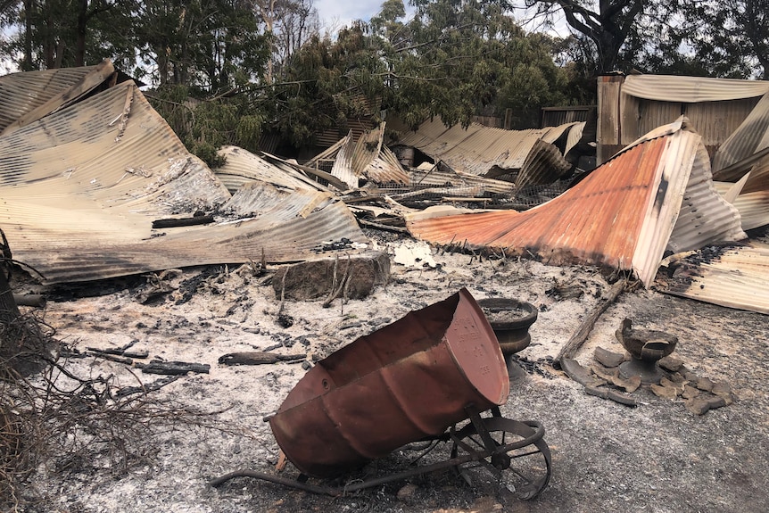 A wide shot of the burnt-out remains of a property destroyed in a bushfire, with an old fashioned wheelbarrow foregrounded.