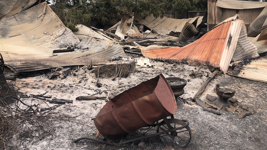 A wide shot of the burnt-out remains of a property destroyed in a bushfire, with an old fashioned wheelbarrow foregrounded.