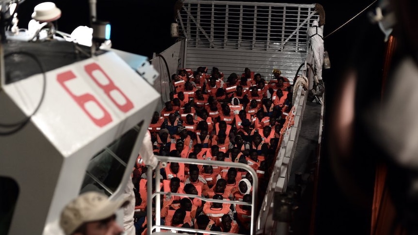 A large group of asylum seekers in life vests sit in a boat after being rescued from the sea.