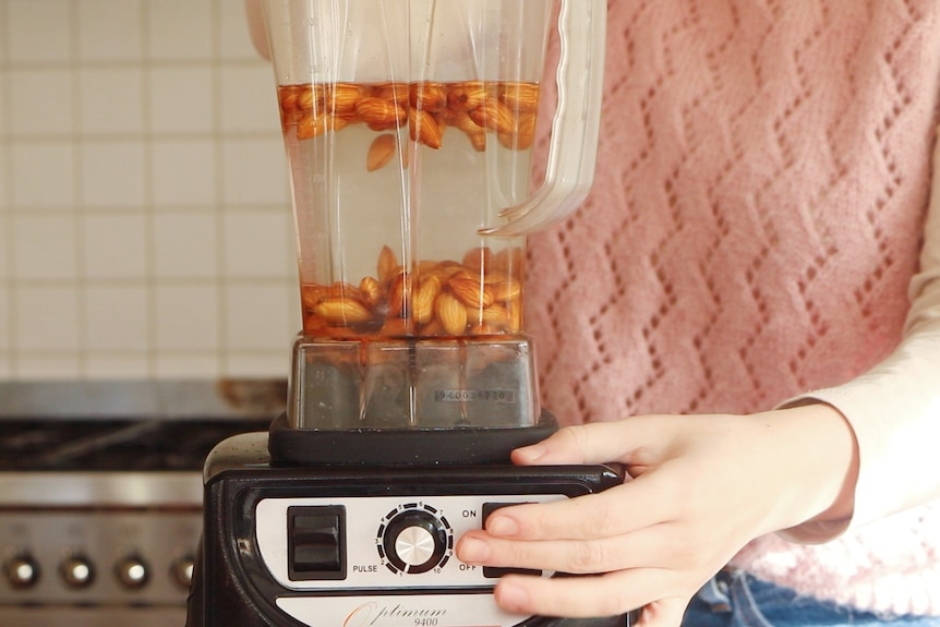 Phoebe is seen with her finger on the on button of a blender containing almonds and water. She stands in a kitchen.