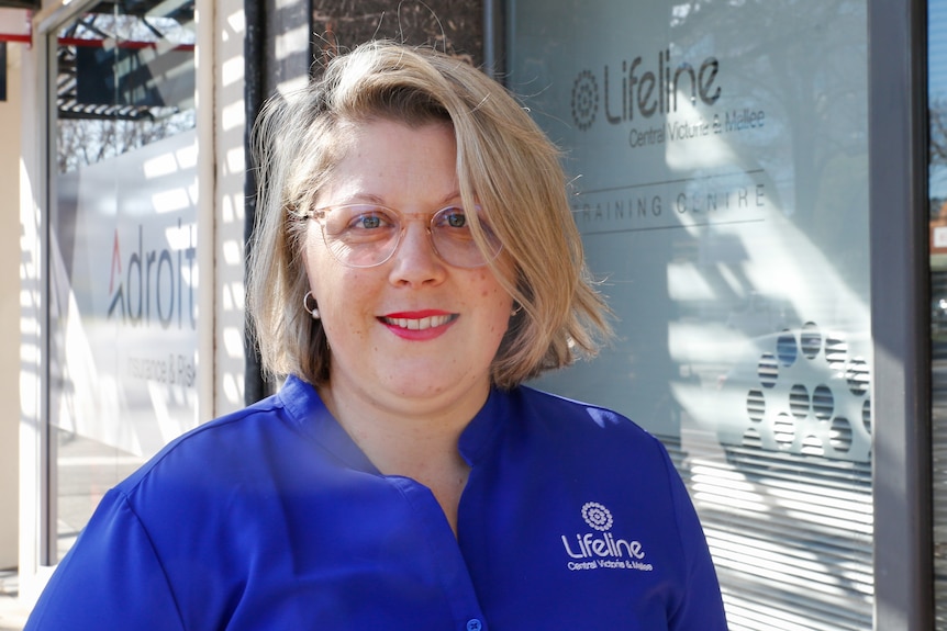 A blonde woman in glasses, blue shirt stands outside a Lifeline centre, smiling slighty.