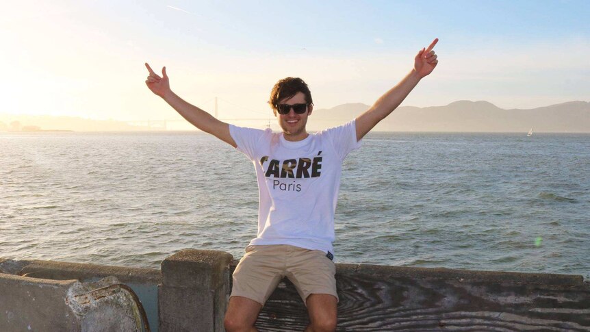 A young man in sunglasses sits by the ocean, smiling, his arms in the air.