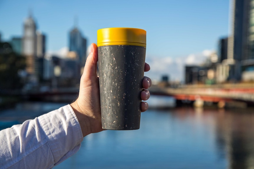 A cup is seen with the city in the background, and a hand holding it. The sky is blue and the river under it is too.
