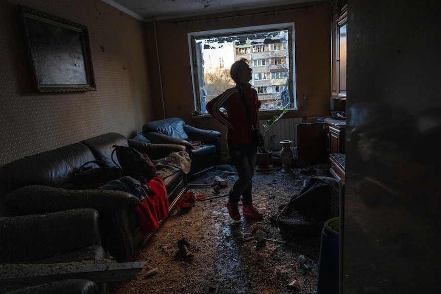 A woman looks around the destroyed living room of her flat, with shattered glass and wreckage strewn about.
