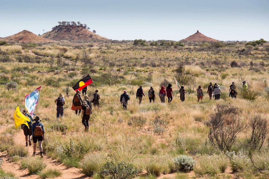 About 25 people in a line march through outback scrubland, some holding flags.