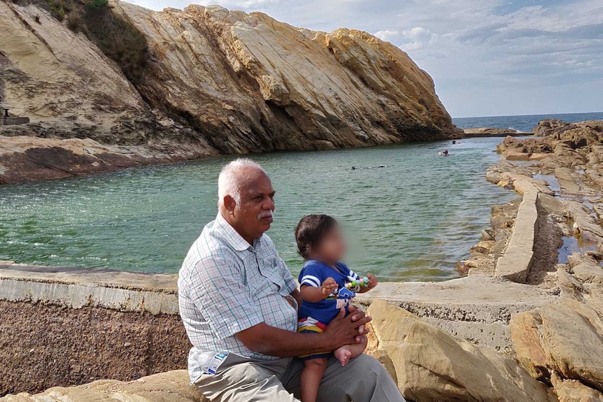 Srihari's dad sitting by an inlet and rocks near the sea, with his grandson on his lap