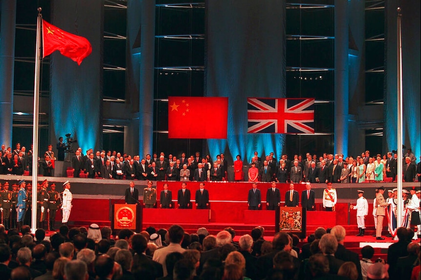 Among a crowd, you look up at a stage with Chinese and British dignitaries as British soldiers lower the Union Jack.