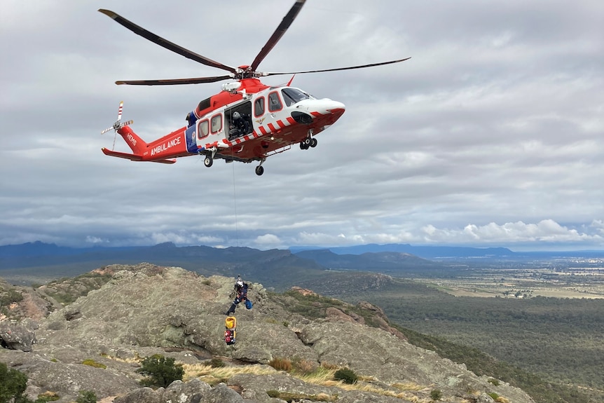 An air ambulance red helicopter hovers over a mountain top waiting for rescue crews to secure a patient. 