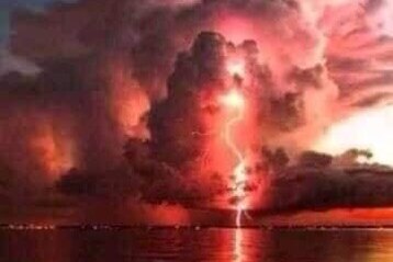 A photo of a red sky and a huge lightning bolt with smoke and clouds over water
