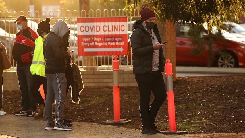 A man looks at his phone with headphones and a face mask on while in a queue outside a COVID testing clinic.