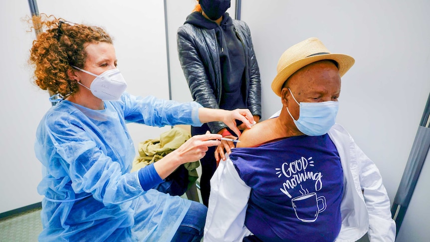 A red-headed nurse in a face mask injects a needle into the arm of a man wearing a boater hat