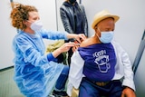 A red-headed nurse in a face mask injects a needle into the arm of a man wearing a boater hat