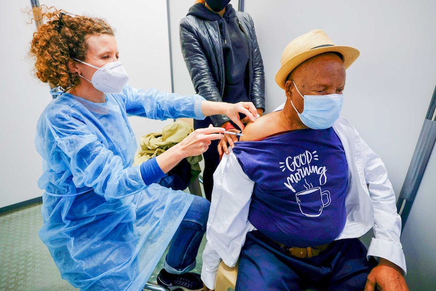 A red-headed nurse in a face mask injects a needle into the arm of a man wearing a boater hat