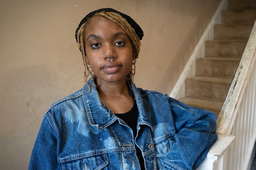 A young woman in a denim jacket leans on the railing of the staircase in her home