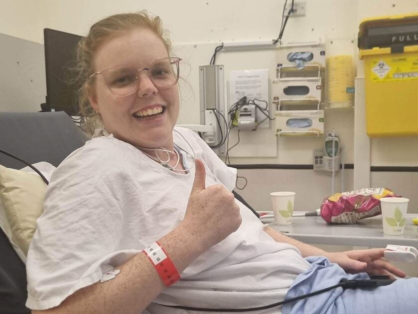A woman with orange hair and glasses gives a thumbs-up to the camera from a hospital bed.