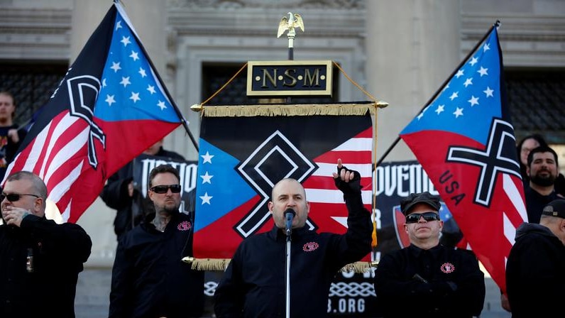 Jeff Schoep speaking into a microphone with neo-Nazi flags in the background.
