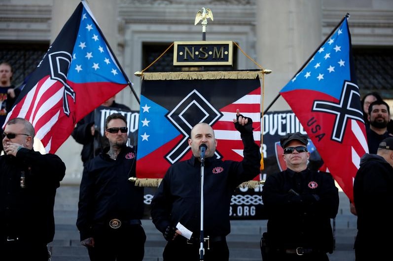 Jeff Schoep speaking into a microphone with neo-Nazi flags in the background.